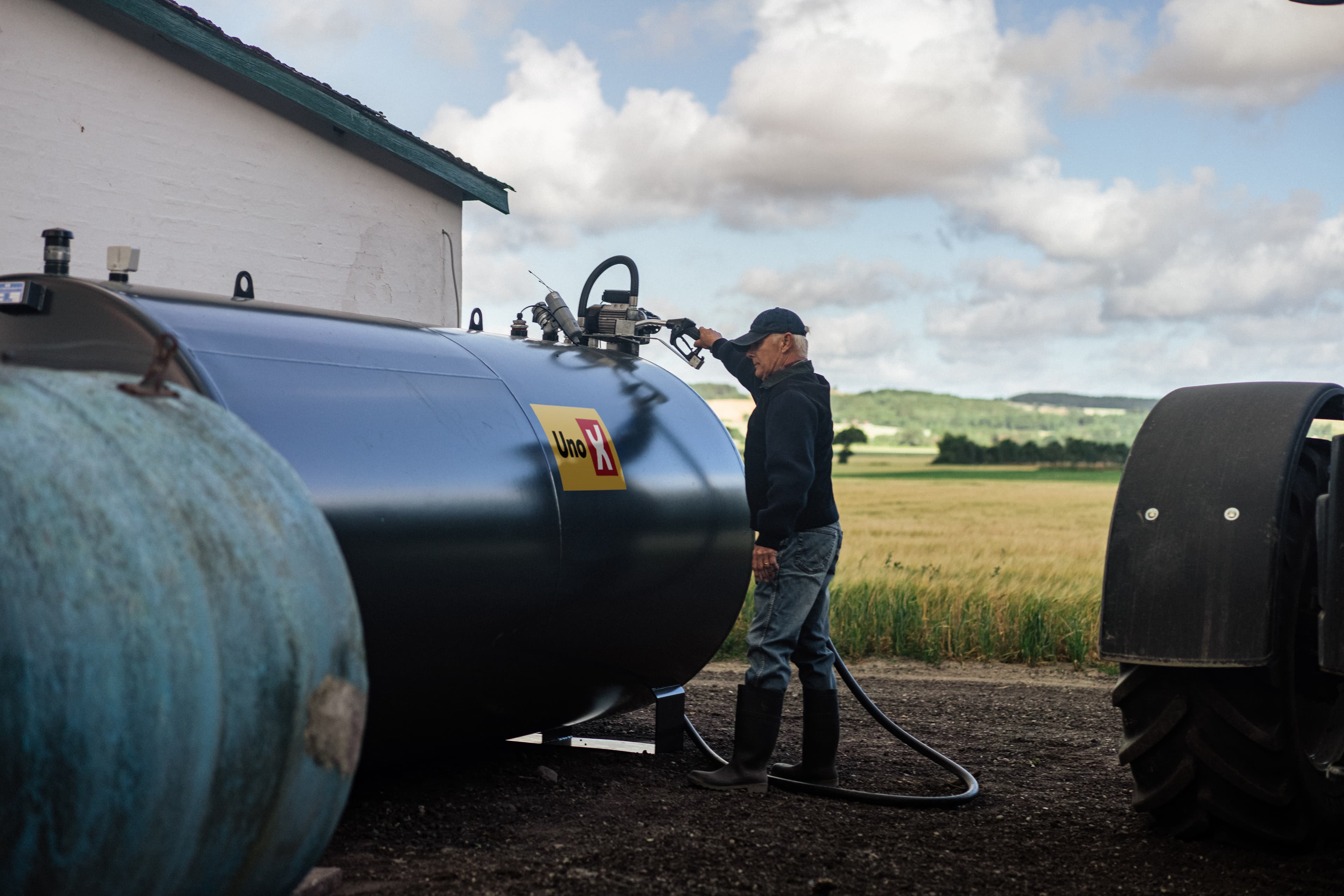 En landmand fylder diesel fra en tank med Uno-X logo på en gård, omgivet af landlige marker og landskab.